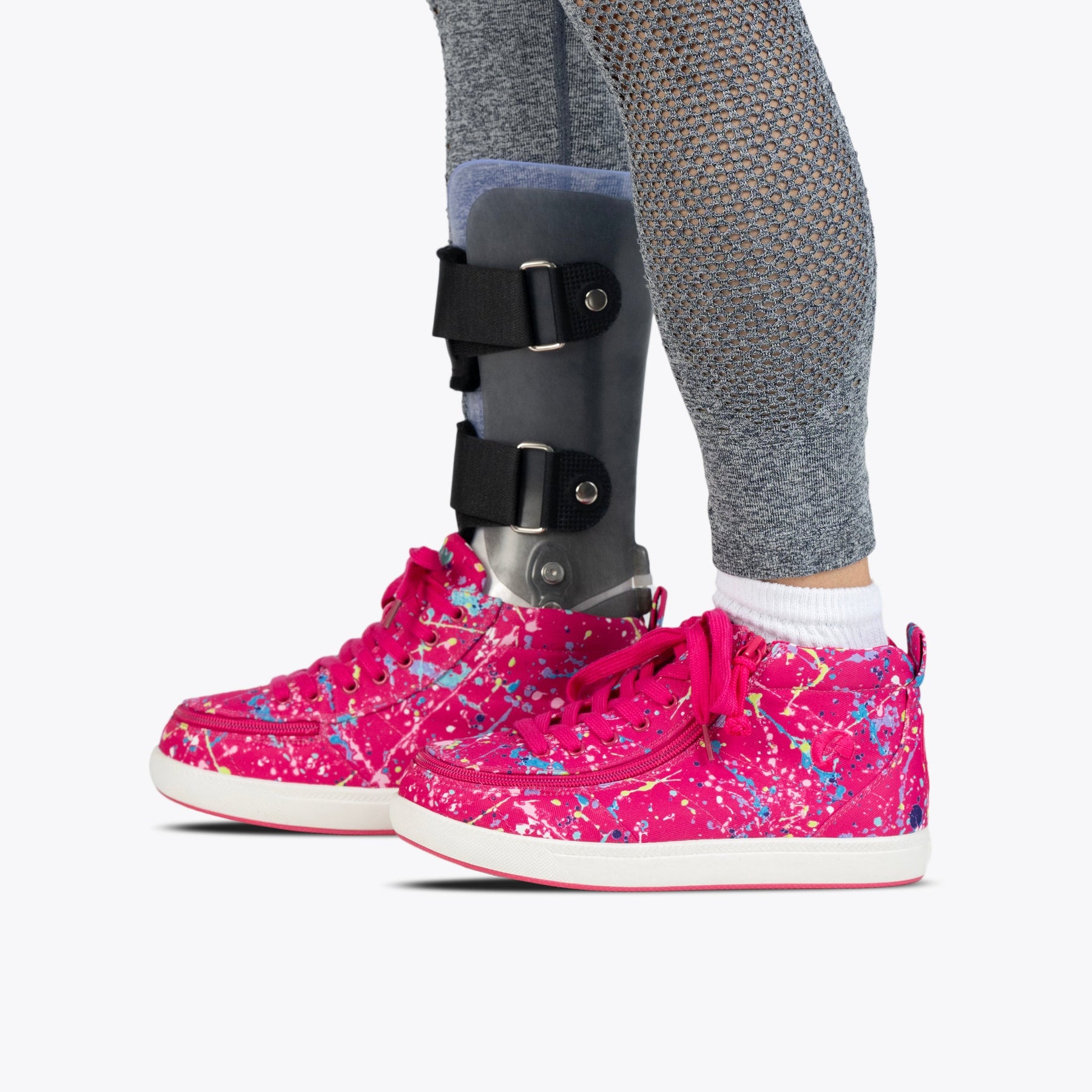 A person wearing grey leggings and an orthopedic leg brace stands on a white background, sporting BILLY Footwear’s Sasha Splatter BILLY Classic D|R II High AFO-friendly shoes in bright pink with a paint-splatter design.