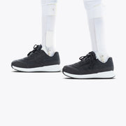 A person with prosthetic legs wears Black BILLY Goat orthotic shoes by BILLY Footwear, featuring white soles, standing on a plain white background.
