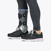 A person wears dark jeans and black sneakers—Midnight Mermaid BILLY Goat by BILLY Footwear—with pastel soles and pink laces, standing with a black leg brace on the left leg. The AFO-friendly shoes support orthotic needs, and the brace has straps and buckles.