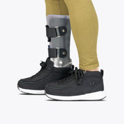 A person wearing gold leggings, a prosthetic leg with metal supports, and Black BILLY Goat Classic High shoes from BILLY Footwear stands against a plain white background.