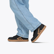 A person steps forward against a plain light background, wearing light blue jeans and Black/Gum BILLY CS High sneakers from BILLY Footwear with a stylized tan E logo.