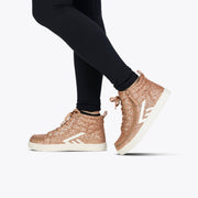 A person in black leggings and BILLY Footwear's Rose Gold Glitz BILLY CS High kids' sneakers with white soles and chevron accents stands on a white background.