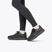 A person wearing Black to the Floor BILLY Sport Inclusion shoes from BILLY Footwear and black leggings stands on a light gray background, with only their legs and feet visible.