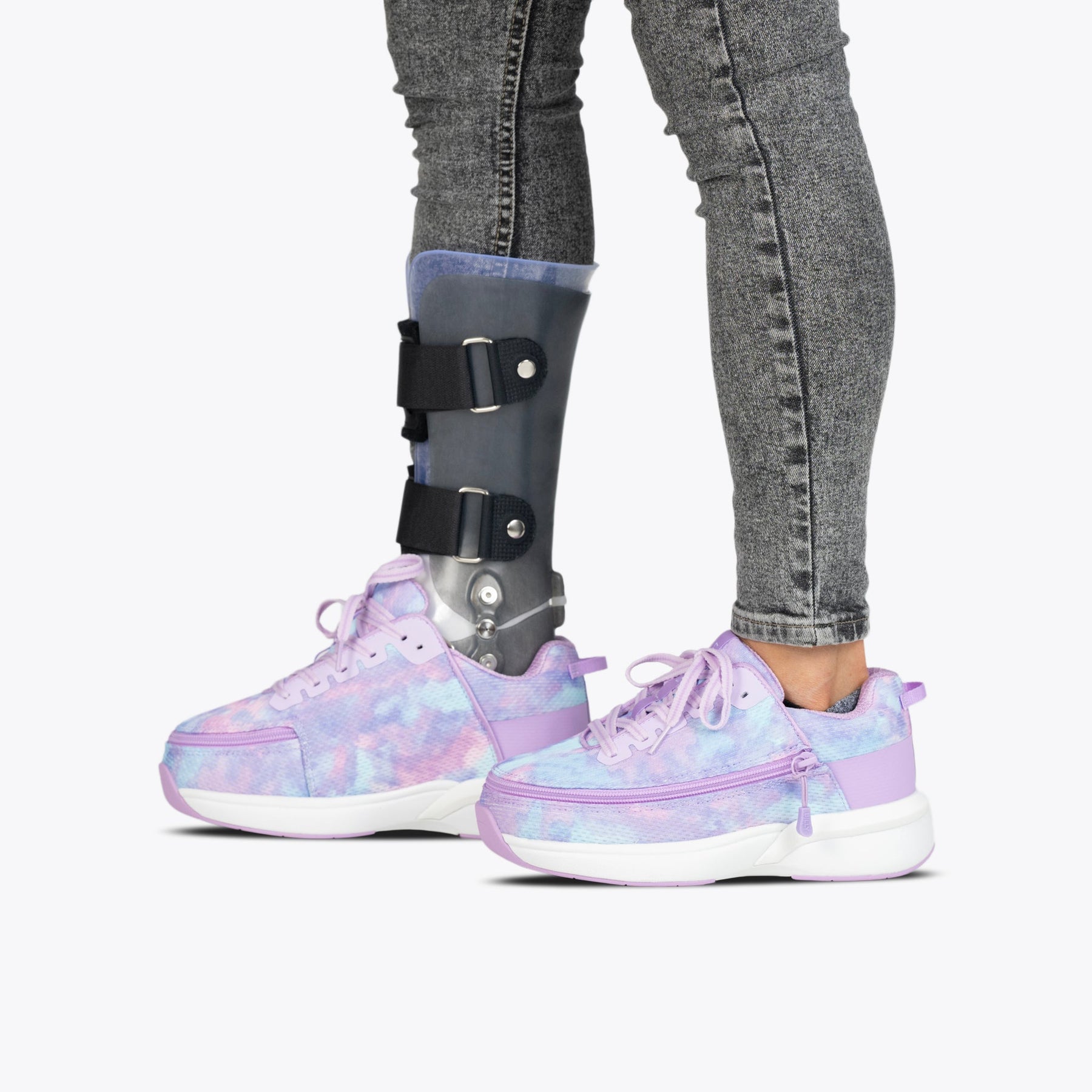 A person wearing gray jeans and mismatched BILLY Footwear Lilac Watercolor BILLY Limitless adaptive shoes, with a visible prosthetic leg brace on the left leg, stands against a plain white background.