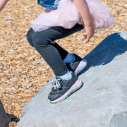 A child in black leggings, a pink tutu, and BILLY Footwear's Midnight Mermaid BILLY Goat sneakers with pastel soles climbs a large gray rock at a wood chip-covered playground.