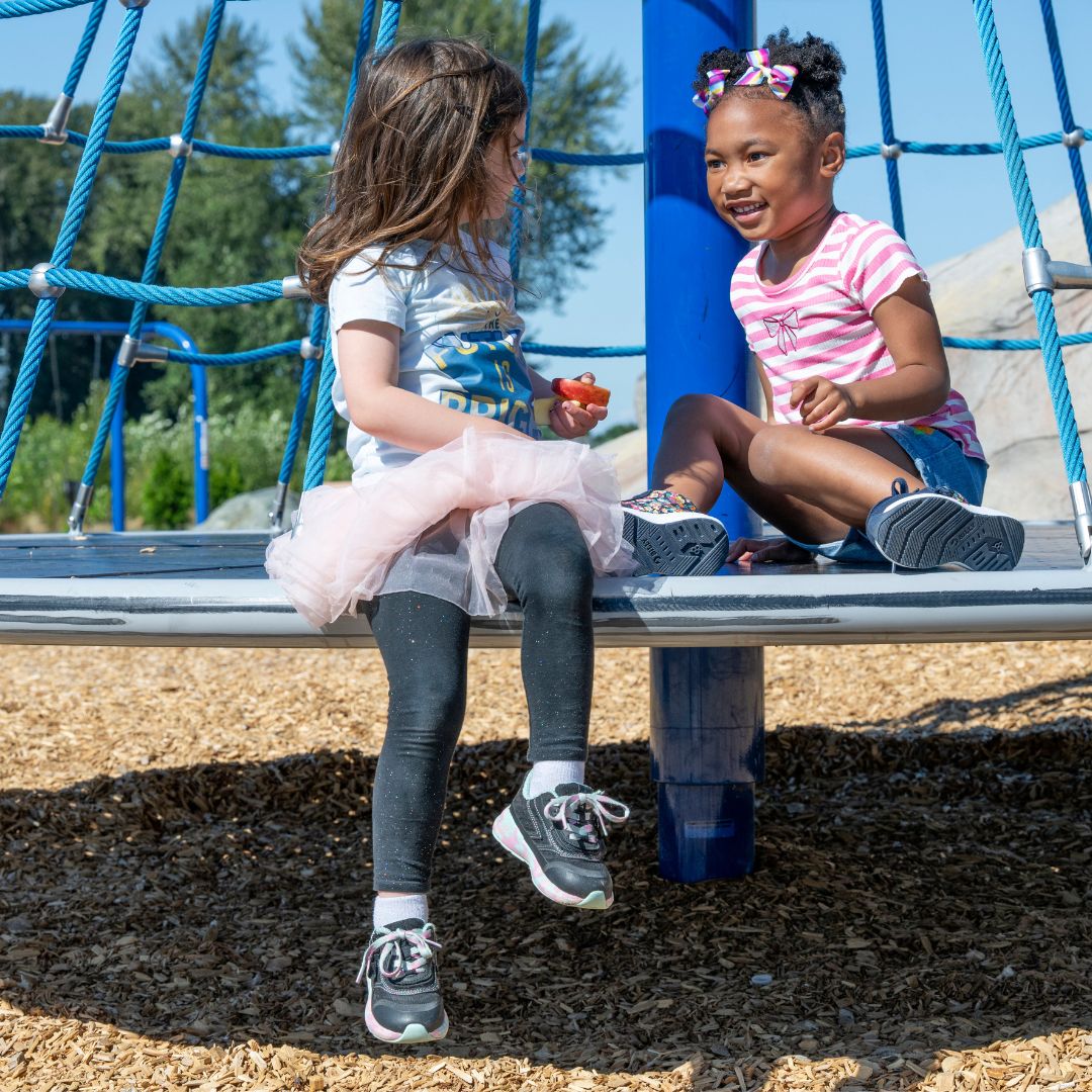 Two young girls smile and chat on a playground climber. One wears a tutu, leggings, and BILLY Footwear's Midnight Mermaid BILLY Goat shoes, designed to be AFO-friendly. The other sports a striped shirt with bows in her hair. Wood chips cover the ground below.