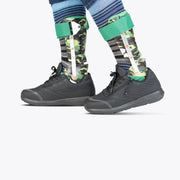 Two lower legs wearing BILLY Footwear Men's Black to the Floor BILLY Goat shoes and camouflage-patterned orthotic leg braces with green straps, over striped socks and blue clothing, set against a plain white background.