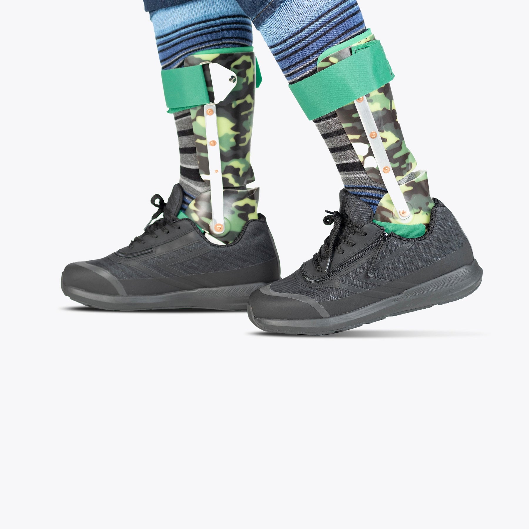 Two lower legs wearing BILLY Footwear Men's Black to the Floor BILLY Goat shoes and camouflage-patterned orthotic leg braces with green straps, over striped socks and blue clothing, set against a plain white background.