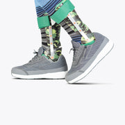 Two prosthetic legs with camouflage-pattern covers and striped socks are shown wearing BILLY Footwear's Men's Charcoal BILLY Goat shoes. The focus is on the lower legs and feet against a plain white background.