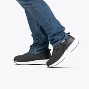 A person is shown mid-step against a plain white background, wearing blue jeans and Men's Black BILLY Limitless shoes with white soles by BILLY Footwear.