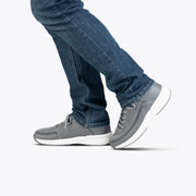 A person in blue jeans wears BILLY Footwear Men's Charcoal BILLY Limitless shoes with white soles, stepping forward on a plain white background.