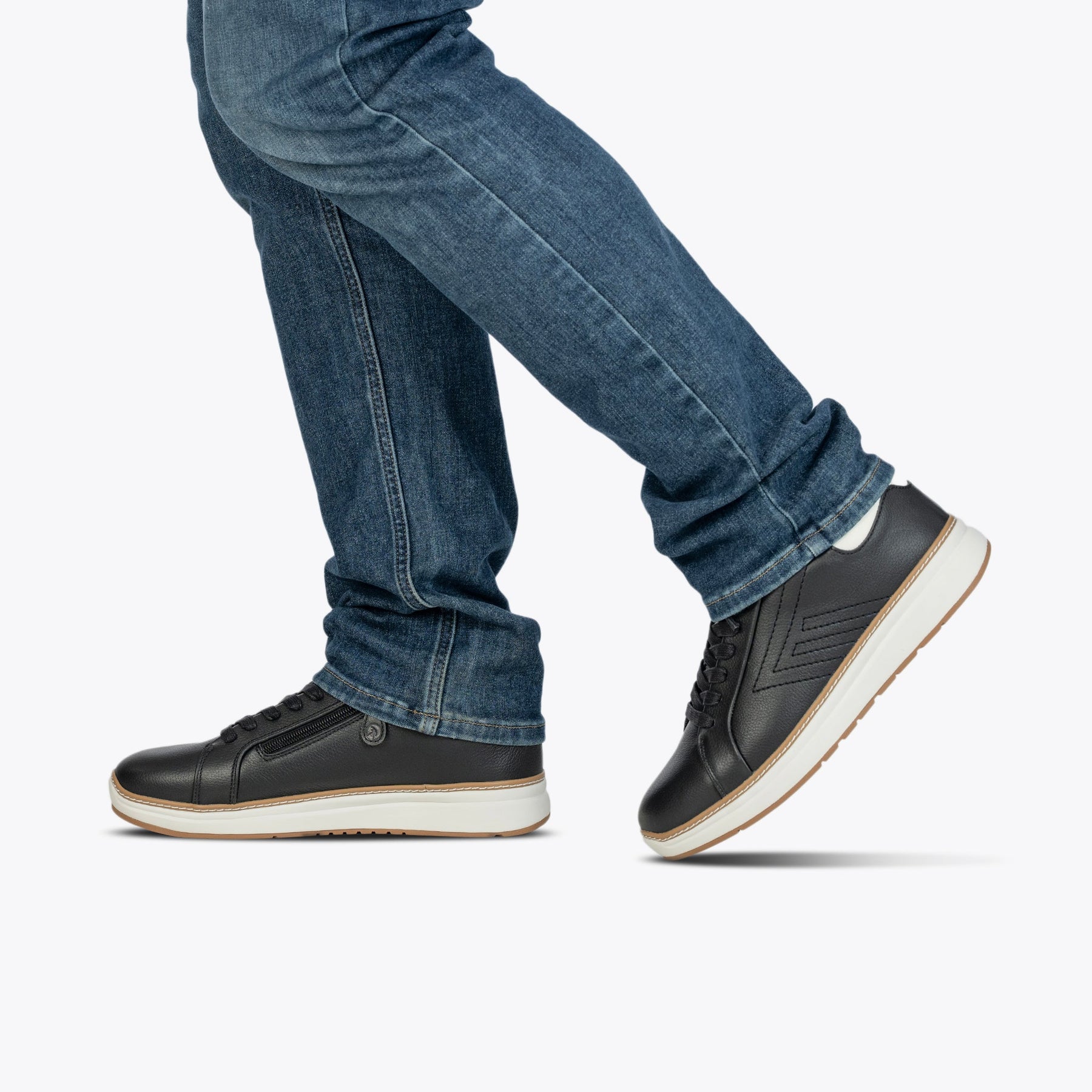 A person in blue jeans and Men's Black Leather BILLY Comfort Luxe sneakers by BILLY Footwear is captured mid-step against a plain white background.