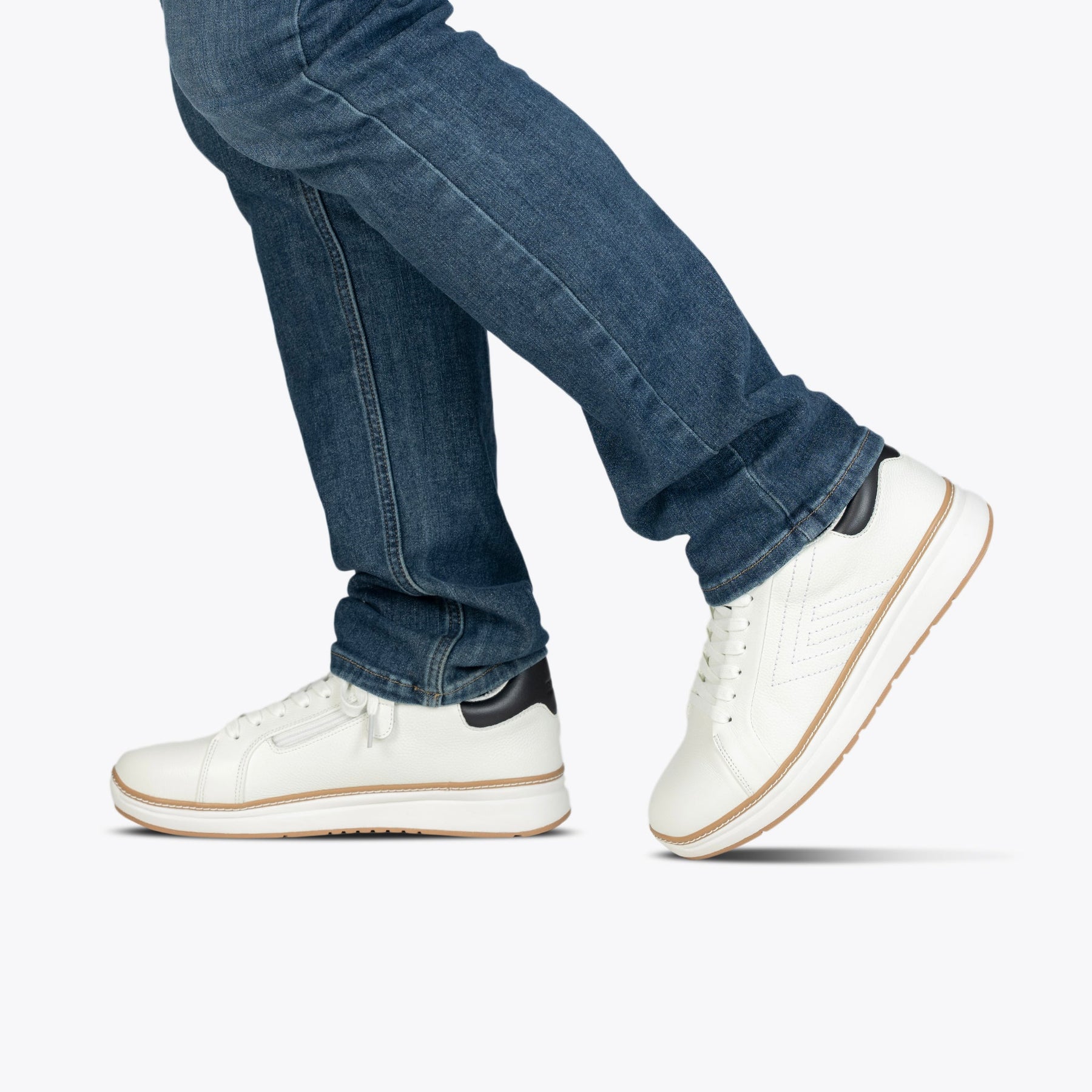 A person in blue jeans wears the Men's White Leather BILLY Comfort Luxe sneaker by BILLY Footwear, accented with black, standing with one foot slightly raised against a plain white background.