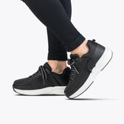 A person is seen from the knees down wearing Women's Black BILLY Limitless shoes by BILLY Footwear, featuring white soles and paired with black leggings, walking against a plain light gray background.